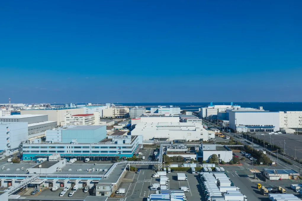 青空と海を背景に広がる、大型倉庫や近代的な工場が集積した広大な臨海工業地域の鳥瞰空撮イメージ。