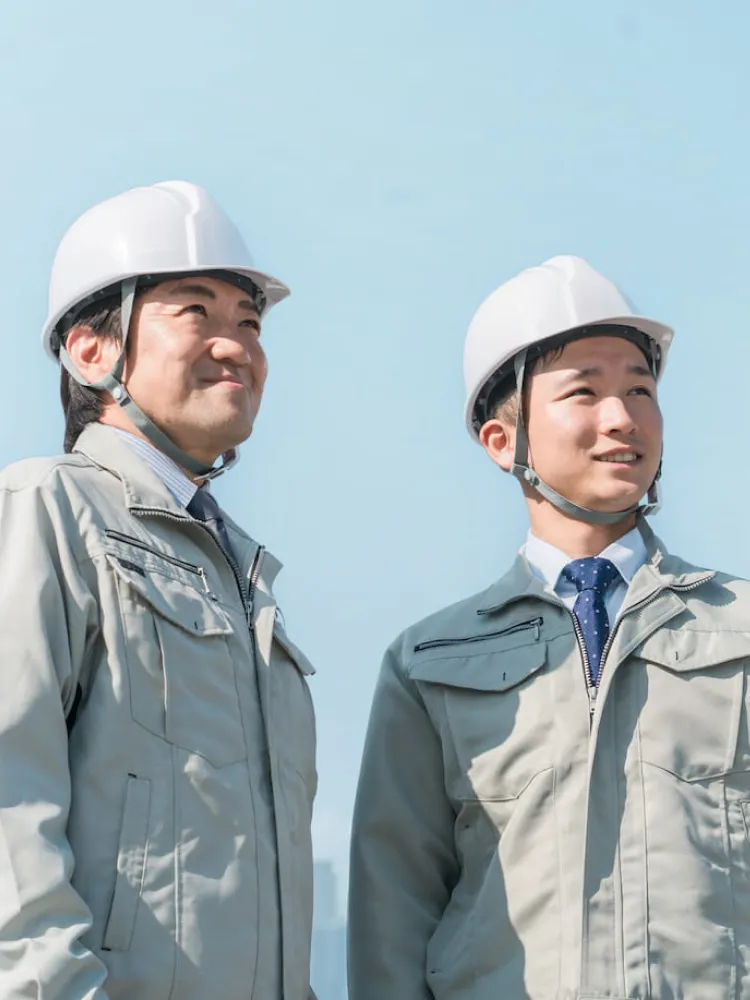 青空の下、白いヘルメットと作業服を着用して前方を明るい表情で見据える二人の男性技術者の様子。