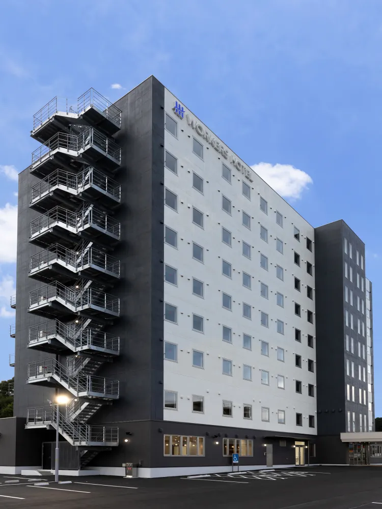 青空を背景に建つ、白とダークグレーを基調としたワーカーズホテルの機能的でモダンな外観と駐車場の様子。