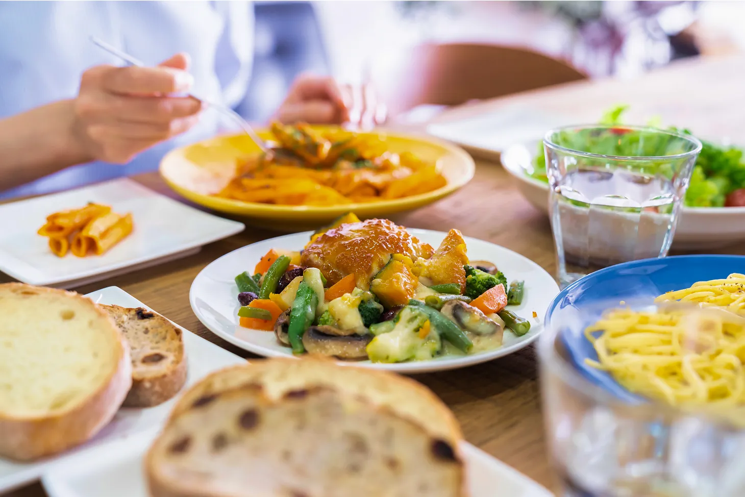 ダイニングテーブルに並べられた、チキンと温野菜のプレート、パスタ、パン、そしてサラダ。手前のチキンと温野菜のプレートには、蒸し鶏のようなメイン料理に、ブロッコリー、にんじん、いんげん、きのこなどのカラフルな野菜が添えられ、食欲をそそります