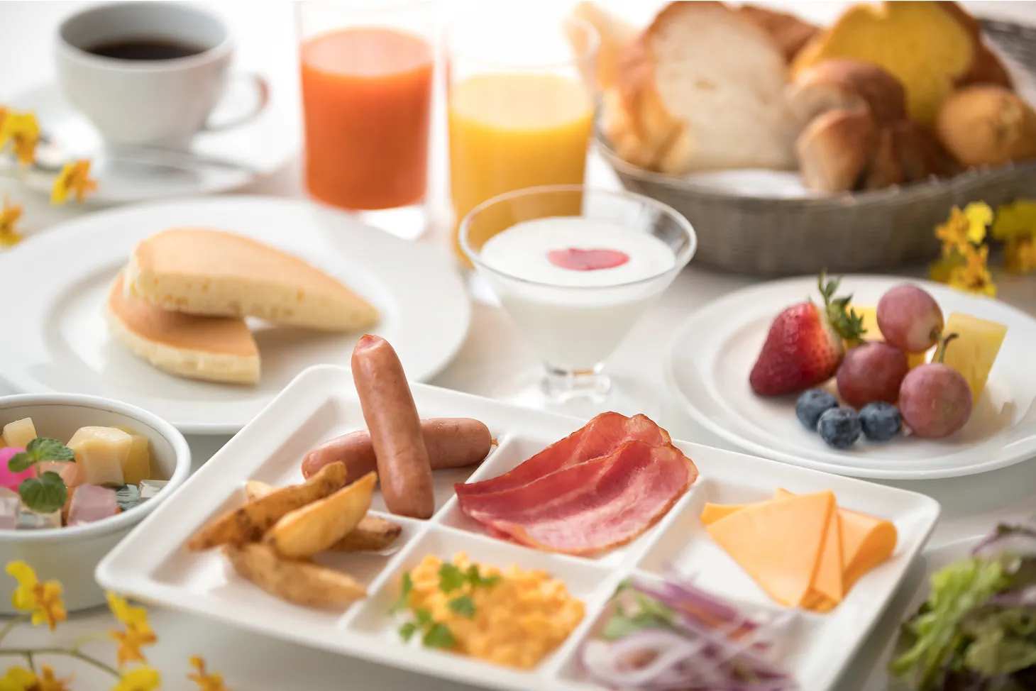 明るい光が差し込むテーブルに並べられた、充実した洋食の朝食ビュッフェ。白い四角いプレートには、ソーセージ、ベーコン、ポテトフライ、スクランブルエッグ、チェダーチーズなどが盛り付けられています。
