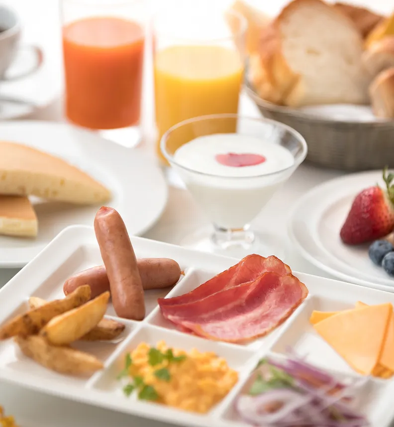 朝食バイキングの料理イメージ。ソーセージ、ベーコン、スクランブルエッグ、チーズなどが盛り付けられた皿や、パンケーキ、ジュースが並んでいる。