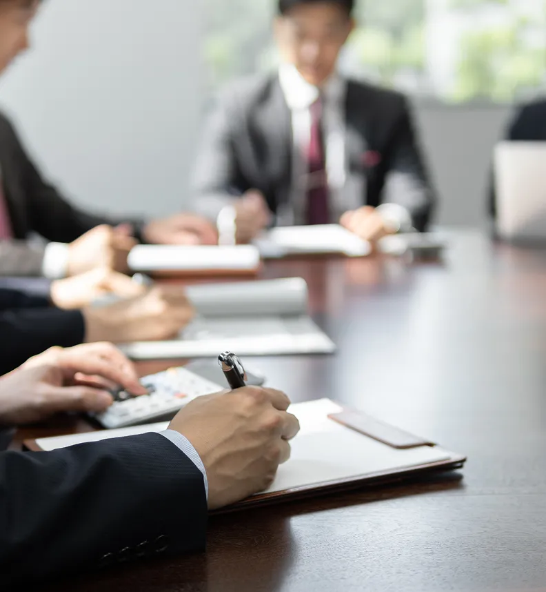 会議スペースの様子。スーツ姿の参加者たちが、大きなテーブルで資料を広げ、熱心に書き込みながら会議をしている。