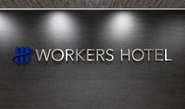 ホテルの客室に置かれた黒いスーツケース。奥には白いベッドとランプが見え、長期滞在に備えている様子。